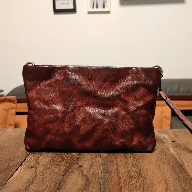 Une pochette en cuir bordeaux posée sur une table en bois de récupération dans un salon, sous une lampe chaude sans lumière d’appoint, un fauteuil orange et un canapé gris derrière